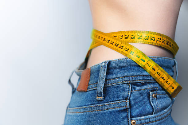 slender young woman measures her slim waist with a tape measure, close-up. Close-up of a slim woman measuring the size of her waist with a tape measure on a white background stock photo