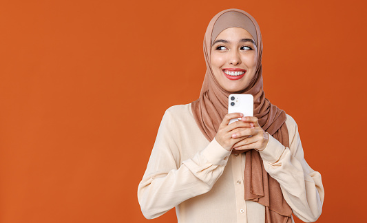 Beautiful overjoyed young smiling muslim woman in traditional religious hijab reading great news on smartphone, amazed young muslim lady receiving unexpected message on mobile phone, isolated on orange studio background