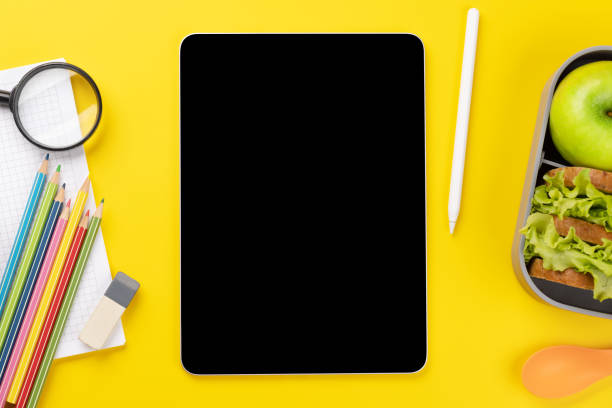 Tablet with blank screen, school supplies, stationery, and lunch box on yellow background. Education and nutrition. Flat lay with blank space for your text or app