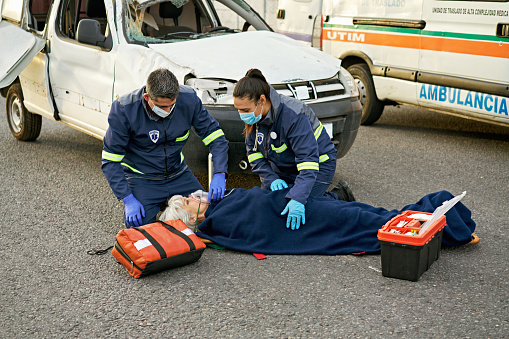 Full length view of uniformed emergency team comforting mature woman lying on stretcher in middle of urban road, damaged vehicle and ambulance in background.
