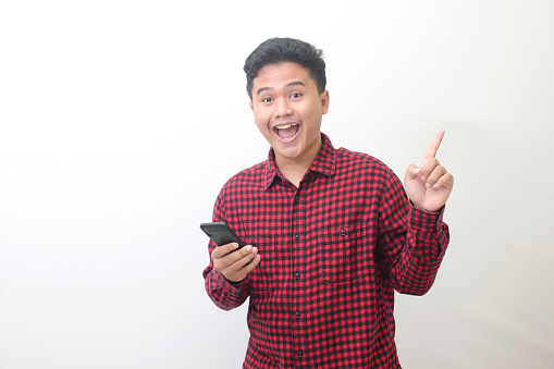 Portrait of attractive Asian man in casual plaid shirt standing against gray background, pointing away with his finger while using mobile phone. Advertising concept.