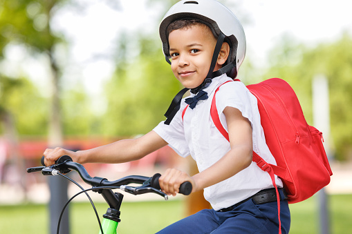 Happy african american child schoolboy in a helmet and with a briefcase rides a bike to school