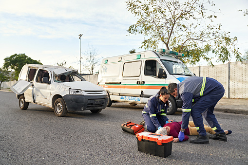 Full length view of ambulance, damaged vehicle, and uniformed man and woman in 20s and 50s attending to woman lying in middle of urban road.
