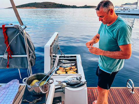 Sailing crew member grilling fishes, potato and cucumbers for dinner on gas grill on sailboat.