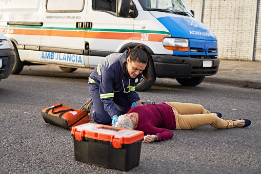 Uniformed woman kneeling next to gray haired woman lying injured in middle of urban road, ambulance in background.