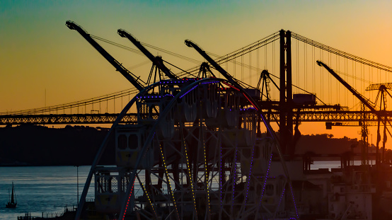 The backlight effect at the end of the day in Lisbon with cranes and dock machines, in the foreground an amusement park wheel