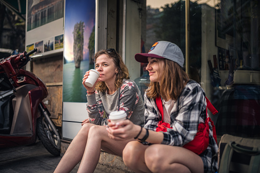 Two young women sitting on the sidewalk and drinking coffee