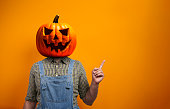 Person in Halloween costume of scarecrow with Jack-o'-lantern pumpkin head points away over orange background with copy space
