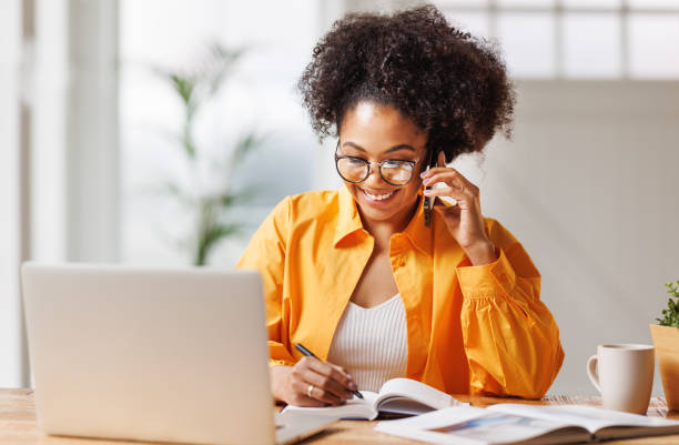 istock Beautiful young smiling ethnic woman making call via smartphone while working remotely from home while sitting at desk and talking to coworkers 1569347962