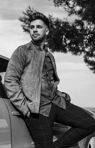 a handsome man leaning on a car parked in a viewpoint while having his hands in the pockets and is looking away | style