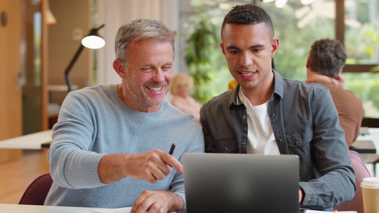 Mature Businessman Mentoring Younger Male Colleague Working On Laptop At Desk