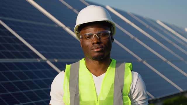Portrait of young handsome African American craftsman in protective helmet and uniform standing among solar panels
