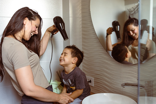 Mother using hairdryer, drying her little son's hair in the bathroom