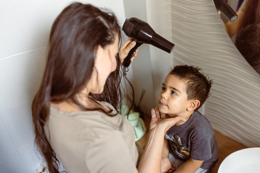 Mother using hairdryer, drying her little son's hair in the bathroom