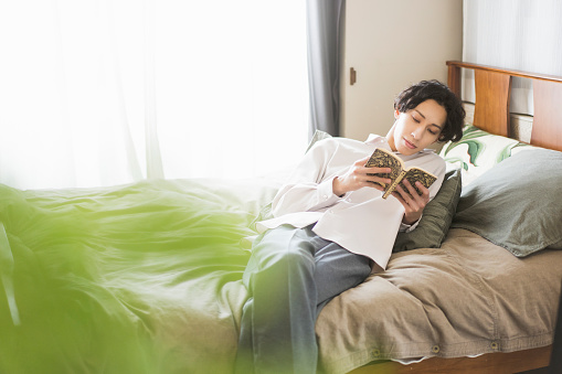 A young asian man is lying on the bed and reading a book.