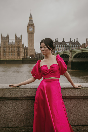 A stunning young Asian fashion model exudes elegance and charm as she strikes a pose on Westminster Bridge, with the magnificent landmarks of Big Ben, Elizabeth Tower, and the Parliament Building creating a breathtaking backdrop. Her pink dress complements the iconic scenery, making this moment a perfect fusion of style and history