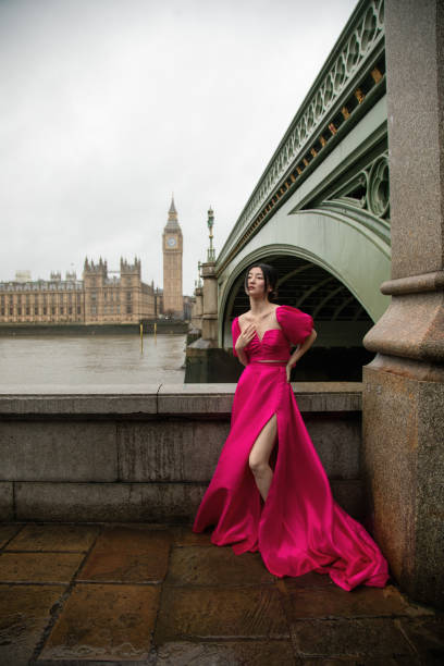 風の強い雨の日にビッグベンと国会議事堂を背景に象徴的なウェストミンスター橋でピンクのドレスを着てポーズをとるスタイリッシュなファッションモデル - elizabeth ii queen nobility british flag ストックフォトと画像