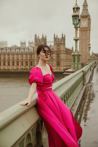 Undeterred by the elements, a stylish young Asian fashion model shines bright in her pink dress on Westminster Bridge. Despite the rainy and windy weather, she gracefully poses in front of the iconic landmarks of Big Ben, Elizabeth Tower, and the Parliament Building. Witness the beauty of resilience and determination as she effortlessly merges fashion and history amidst the atmospheric charm of a blustery day.