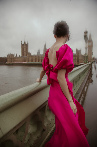 A stunning young Asian fashion model exudes elegance and charm as she strikes a pose on Westminster Bridge, with the magnificent landmarks of Big Ben, Elizabeth Tower, and the Parliament Building creating a breathtaking backdrop. Her pink dress complements the iconic scenery, making this moment a perfect fusion of style and history