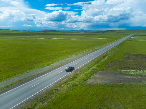 Gray passenger car is driving along an asphalt road along green fields, mountains on a sunny day, beautiful clouds in the sky. Photographing from a drone. Concept of travel