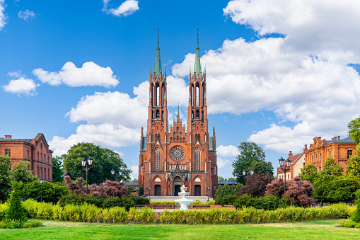 A Gothic-style cathedral located in Żyrardów, Poland. The cathedral is made of red bricks and features two tall towers. Positioned at the center of the photo, it stands against a blue sky with fluffy clouds. In the foreground, there is a neatly trimmed green lawn and bushes. No people are visible in the frame.