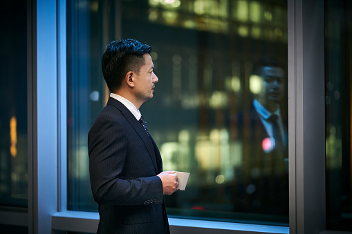 asian corporate executives standing by the window thinking looking at night scene with a cup of coffee in hand