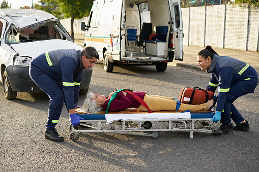 Full length view of male and female emergency team managing mature woman lying on portable gurney next to damaged panel van in urban road.