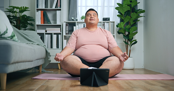 Young Chubby Man Practicing Yoga Exercise with Laptop computer in Living Room, wellness and fitness in harmonious blend of indoor activity, for good health of overweight man