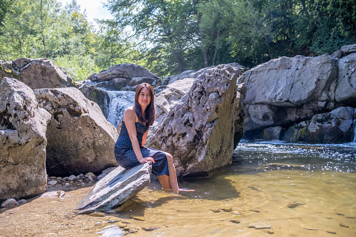 Asian woman sitting on a rock in a river next to a waterfall.