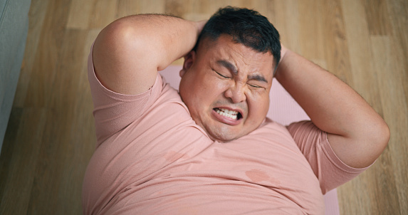Top view, Young chubby man lying on wooden floor in living room trying with difficulty to do sit-up exercises, to reduce the size of his belly, good health of overweight man
