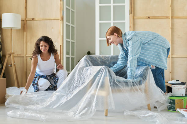 istock Two happy girlfriends wrapping armchair with cellophane packaging 1569780402