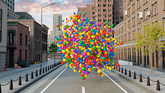 A large collection of colorful balls or balloons flys through the streets of a city. They fly towards the sky and pass by large buildings. It is a surreal sight.