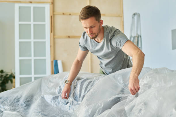 istock Young man in grey t-shirt bending over couch while covering it with cellophane 1569783738