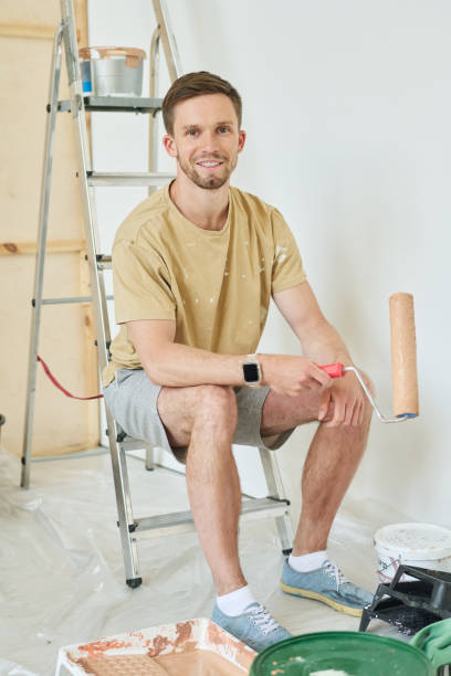 istock Young smiling man with paintroller sitting on stepladder and looking at camera 1569784333
