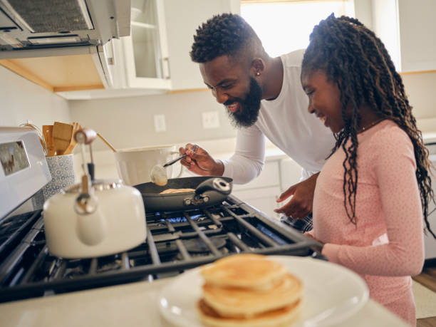 A black father and daughter wearing pajamas, cooking and eating a pancake breakfast together in a home.