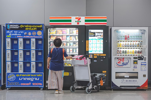 BANGKOK, THAILAND - June 27, 2023 : Female passenger with luggage trolley buying drinks and snack from the 7- eleven vending machines inside of Krung Thep Aphiwat central terminal station building