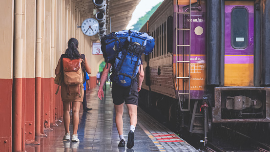 Chiang Mai, Thailand - June 26, 2023 : Rear view of interracial couple travelers with baggage are walking on platform at Chiang Mai train station, full length and wide screen view
