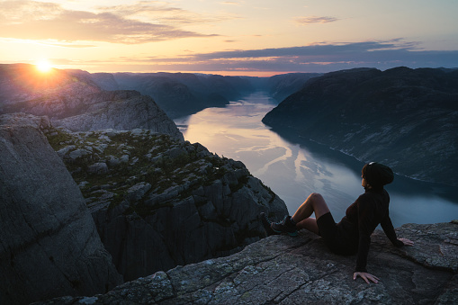 The single person sitting alone on the edge of the cliff Preikestolen in Rogaland county in Norway watching the sun rising. Flat top cliff is a tourist attraction above Lysefjorden fjord. A rare moment of being alone on Pulpit Rock during sunrise in the summer of June 2023. Sun is raising giving the sky and the water beautiful yellow, pink and violet and creating a contrast of warm and cold colours during the golden hour.