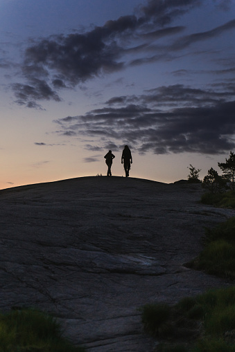 Silhouettes of two people, friends hiking the mountain on the rocky ground towards the rising sun. People going towards Preikestolen in Rogaland county in Norway for the sunrise in the summer of June 2023. Flat top cliff called The Pulpit Rock is a tourist attraction above Lysefjorden fjord. Sun is almost raising giving the sky beautiful yellow, pink and violet colours and creating a contrast of warm and cold colours during the golden hour.