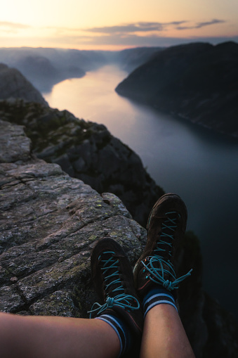 Single person's legs in trekking shoes on the edge of the cliff Preikestolen in Rogaland county in Norway while the sun is rising. Flat top cliff is a tourist attraction above Lysefjorden fjord. A rare moment of being alone on Pulpit Rock during sunrise in the summer of June 2023. Sun is raising giving the sky, water and legs in professional hiking shoes beautiful yellow, pink and violet and creating a contrast of warm and cold colours during the golden hour.