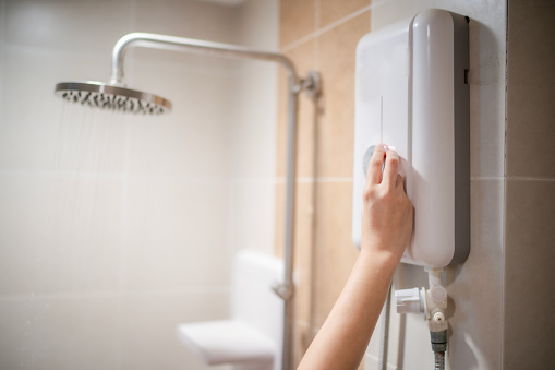 Woman hand adjust temperature at electric water boiler with shower and rain water blur background and space, woman using water boiler in bathroom at home.