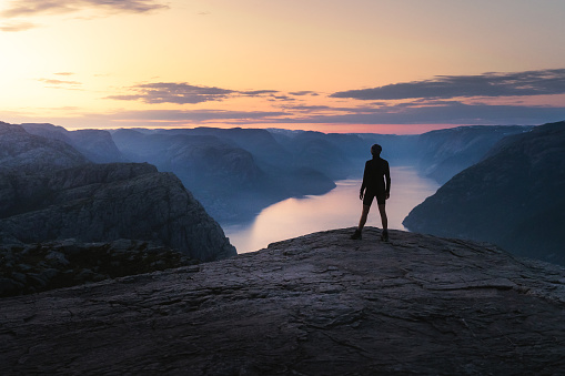 The single person standing alone on the edge of the cliff Preikestolen in Rogaland county in Norway watching the sun rising. Flat top cliff is a tourist attraction above Lysefjorden fjord. A rare moment of being alone on Pulpit Rock during sunrise in the summer of June 2023. Sun is raising giving the sky and the water beautiful yellow, pink and violet and creating a contrast of warm and cold colours during the golden hour.