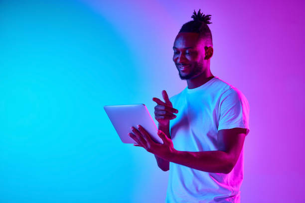 istock Young african man with dreadlocks holding padlet over gradient blue-pink background in neon. 1569844336