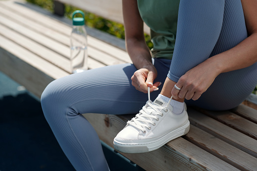 Close-up of young sportswoman in blue leggins tying shoelace of sneaker while sitting on bench after training in open air and having break