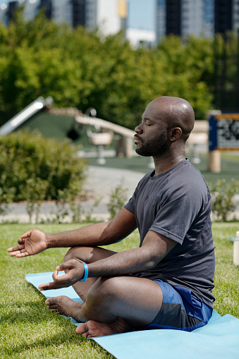 Side view of young African American man in activewear meditating on mat while sitting in pose of lotus during yoga practice in open air