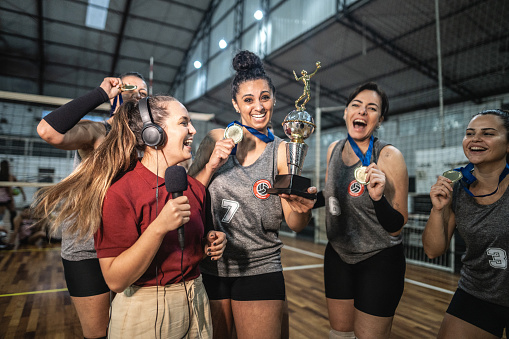 Young woman TV reporter interviewing female volleyball team after win a match on the sports court