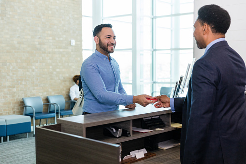 The young adult man smiles as he hands the mature adult male bank teller his debit card.