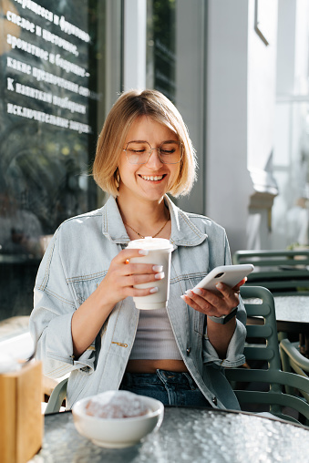 Positive charming laughing young woman with short hair using phone while sitting with cup of coffee in street cafe, summer lifestyle. Cute smiling woman chatting, browsing internet in outdoor cafe.