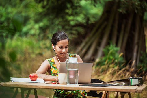 front view on woman sitting at wooden desk on meadow in garden and working at laptop