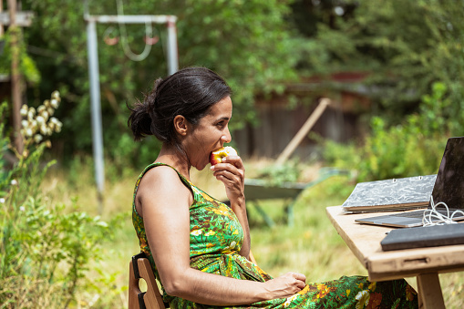 side view on woman sitting at wooden desk with laptop in garden and eating an apple